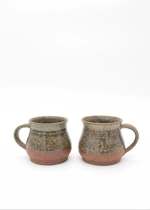 Two handmade ceramic mugs with rustic handles, featuring a speckled glaze of earthy green, brown, and terracotta tones, set against a plain white background.