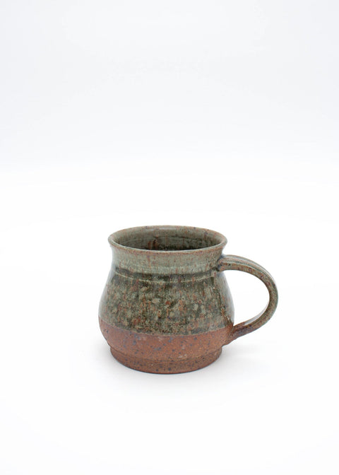 A handmade ceramic mug with a rounded body, a curved handle, and a rustic finish. The upper portion features a speckled green glaze, while the lower portion and base are exposed terracotta clay.