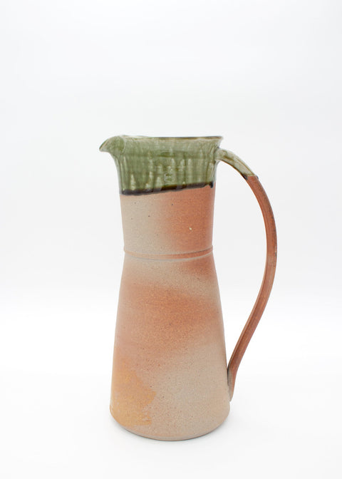 A handmade ceramic pitcher with a warm terracotta orange body, a textured green glazed rim, and a curved brown handle, set against a plain white background.